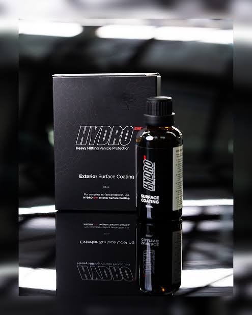 A bottle of HYDRO9G+ Exterior Surface Coating next to its black packaging box, displayed on a reflective dark surface with a blurred automotive background.